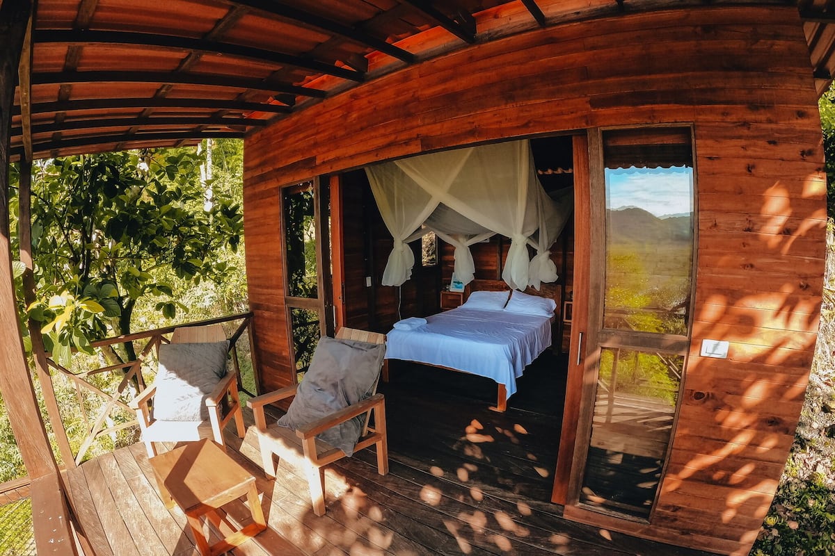 Eine Holzhütte mit einem Bett, zwei Stühlen und einem Tisch auf einer Veranda.
