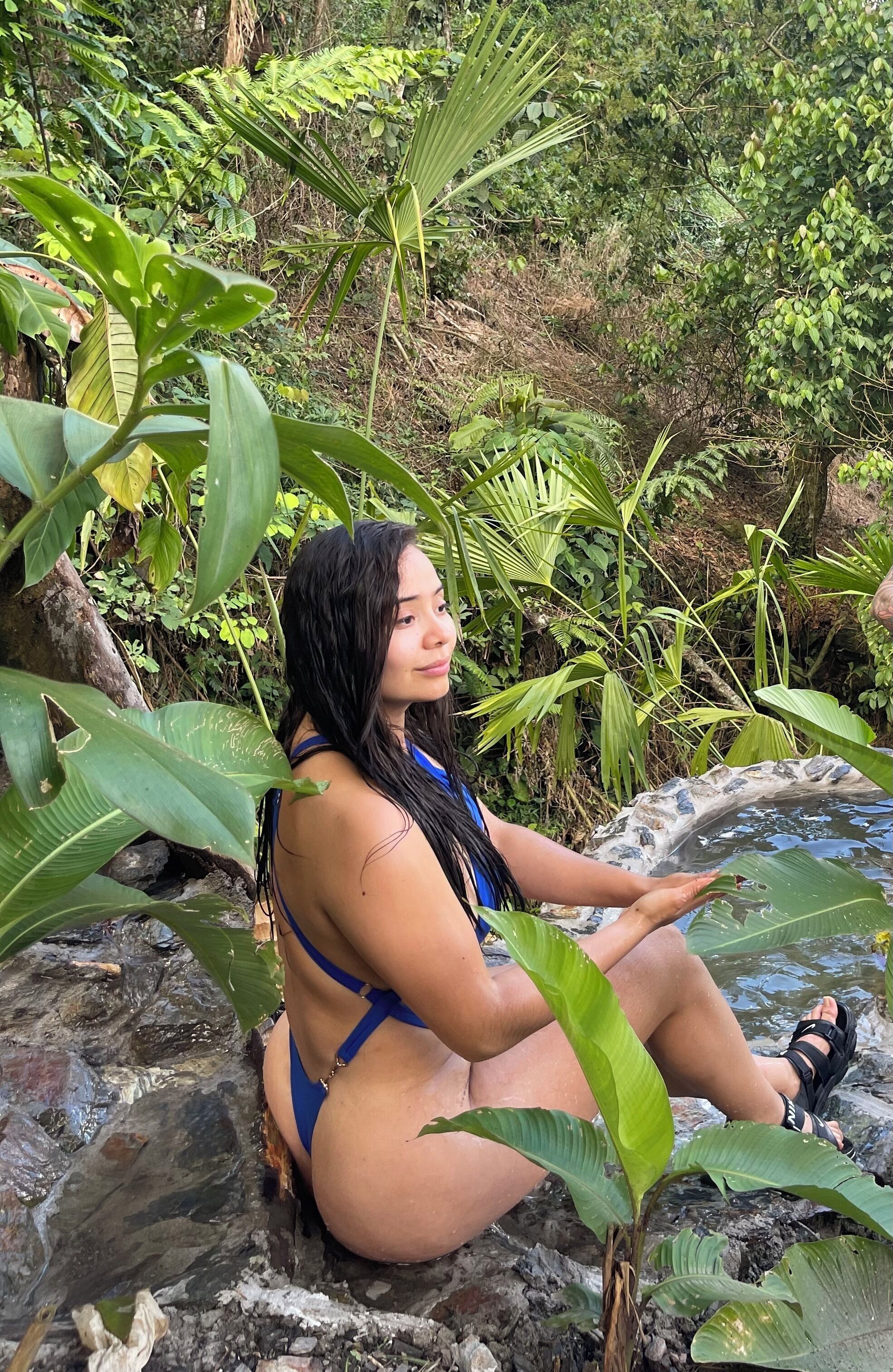 Una persona sentada sobre rocas junto a una fuente de agua, rodeada de abundante vegetación.