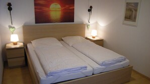 A double bed with two pillows, a nightstand with a lamp, and a painting of a sunset on the wall. - Bungalow 21 on the edge of the forest with terrace and view of the Wasserkuppe. (Dipperz)