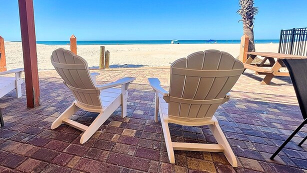 A beachside patio with Adirondack chairs and a wooden table, overlooking a sandy beach and clear blue ocean.