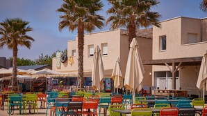An outdoor seating area with colorful chairs and umbrellas, palm trees, and a building with windows and a mural. - ⛱️ Direct beach access - 4* campsite All-comfort mobile home + Covered terrace (Vias)