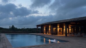 A swimming pool with a wooden deck and lounge chairs, surrounded by a building with large windows and a covered patio area. - Villa Campo Verde – Luxury, nature & relaxation in Provence (saint martin de crau)