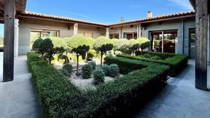 Une cour dotée d’un jardin à la disposition symétrique, avec des haies et des arbres taillés avec soin, et un bâtiment surmonté d’un toit en tuiles et doté de grandes fenêtres.