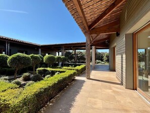Ein überdachter Patio-Bereich mit Fliesenboden, umgeben von ordentlich geschnittenen Hecken und Bäumen.