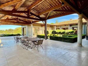 Eine überdachte Terrasse mit Esstisch und Stühlen, Steinboden und Blick auf ein Haus mit Ziegeldach und gepflegtem Garten.