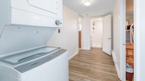 Un cuarto de lavandería con lavadora y secadora de carga frontal, piso de madera y un pasillo que conduce a una habitación con puerta.
