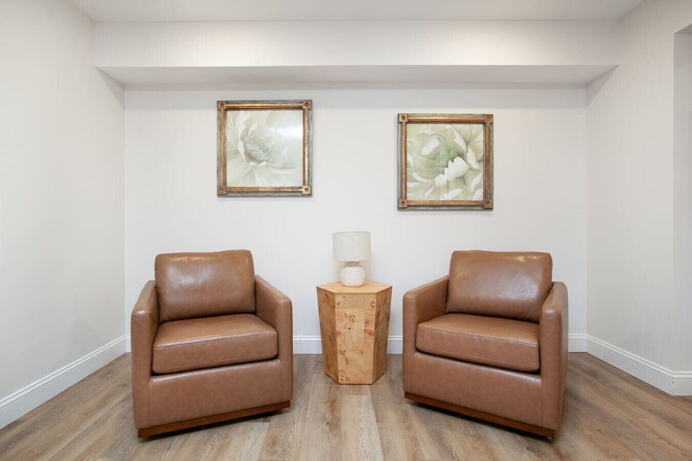 A room with two leather chairs, a wooden side table with a lamp, and two framed abstract paintings on the wall.