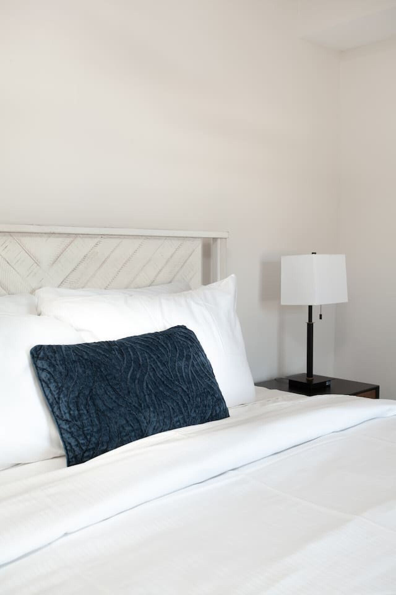 Een netjes opgemaakt bed met witte lakens en een donkerblauwe kussen, een wit hoofdbord en een nachtkastje met een lamp.