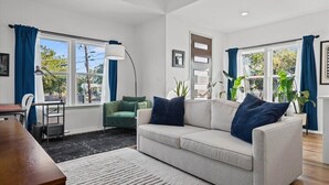 A modern living room with a sofa, a desk, and a chair. There are large windows with curtains, a floor lamp, and potted plants.