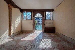 Ein Korridor mit Schachbrettboden, einer Holztür mit Glasfeldern und Blick auf ein Gebäude hindurch.