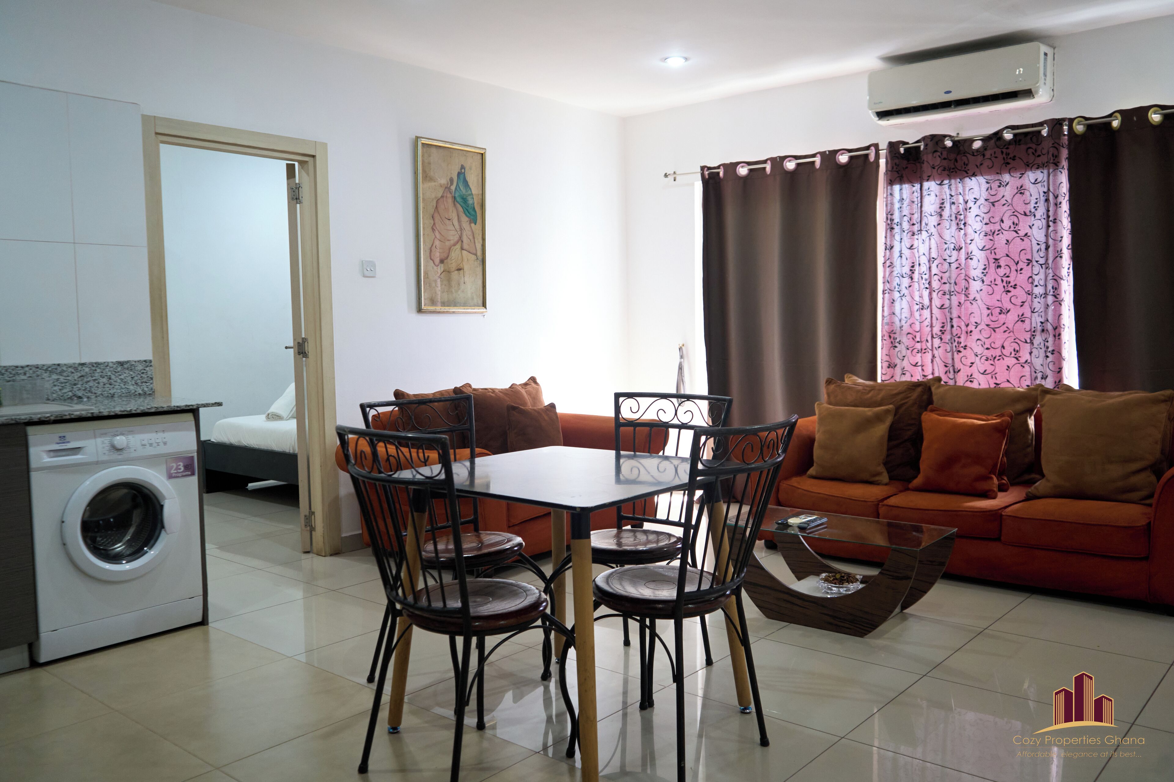 A compact living space with a dining area, a sofa, a washing machine, and a window with curtains.