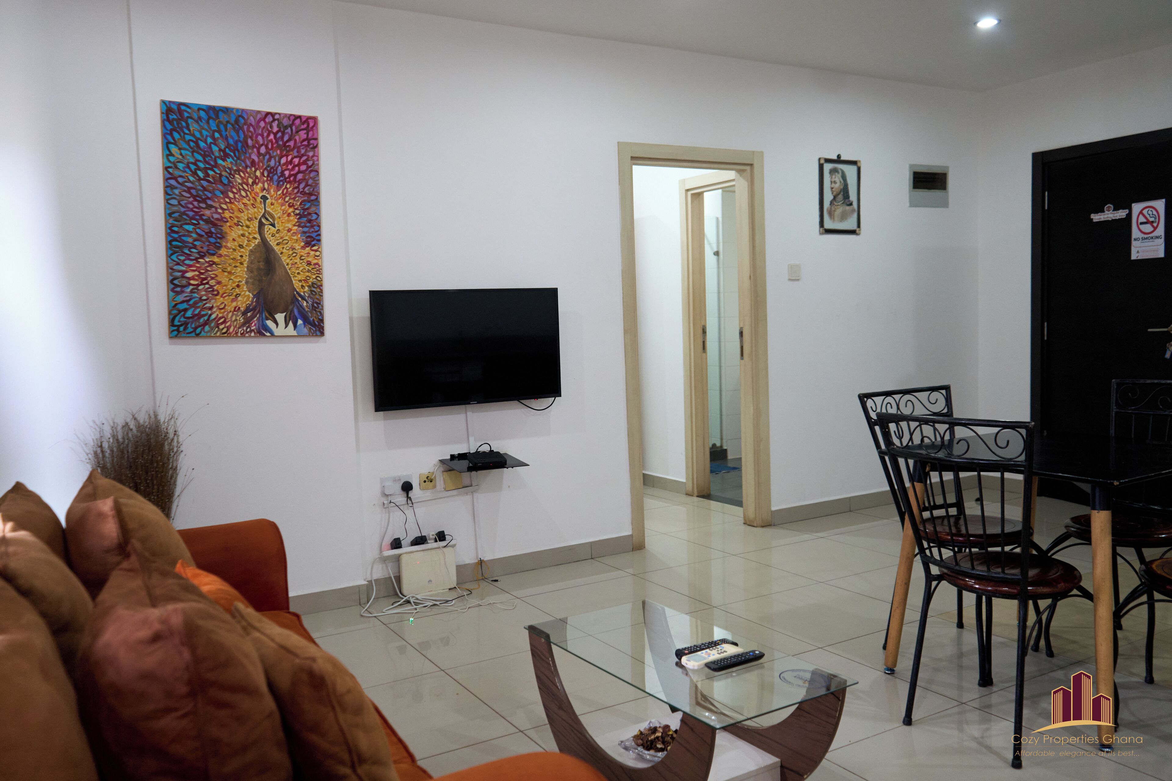 Ein Wohnzimmer mit Flachbildfernseher, Glastisch, Esstisch mit Stühlen und einem farbenfrohen Pfauenbild an der Wand.