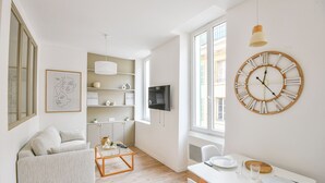 A modern living room with a sofa, coffee table, and dining area with a clock and TV. - Bail mobilité - Appart standing proche Vieux-Port (Marseille)