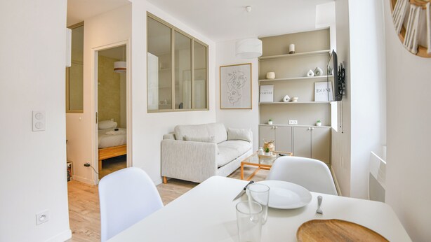 A modern, well-lit dining area with a white table set for two, a sofa, and a bookshelf with various items. - Bail mobilité - Appart standing proche Vieux-Port (Marseille)
