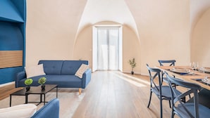 Un salon moderne avec un canapé bleu, une table à manger dressée pour quatre personnes et une grande fenêtre en arc de cercle.