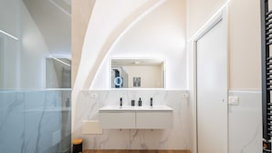 Une salle de bain moderne avec un mur en marbre, un plancher en bois, un meuble-lavabo blanc avec un miroir et une poubelle noire.