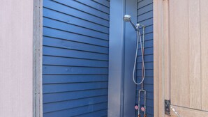 Un petit espace de douche extérieur, à l’abri, doté d’une porte en bois et d’un pommeau de douche en métal.