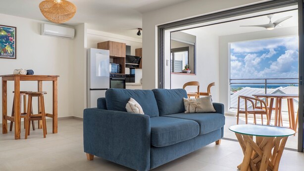 Ein modernes Wohnzimmer mit einem blauen Sofa, Holzstühlen, einem Glastisch und einer Küche im Hintergrund.