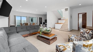 A spacious living room with a sectional sofa, a coffee table with a decorative bowl, and patterned armchairs. A staircase with a glass railing leads to an upper level. - Casa Oasis B-Nayar Gated Beach Club (La Cruz de Huanacaxtle)