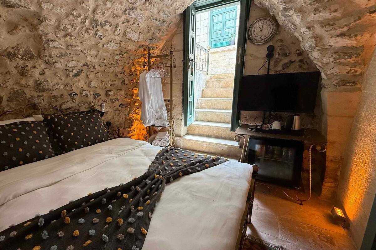 Une chambre rustique avec un mur en pierre, un lit agrémenté d’oreillers à motifs, une petite table avec une horloge et une fenêtre donnant sur l’extérieur.