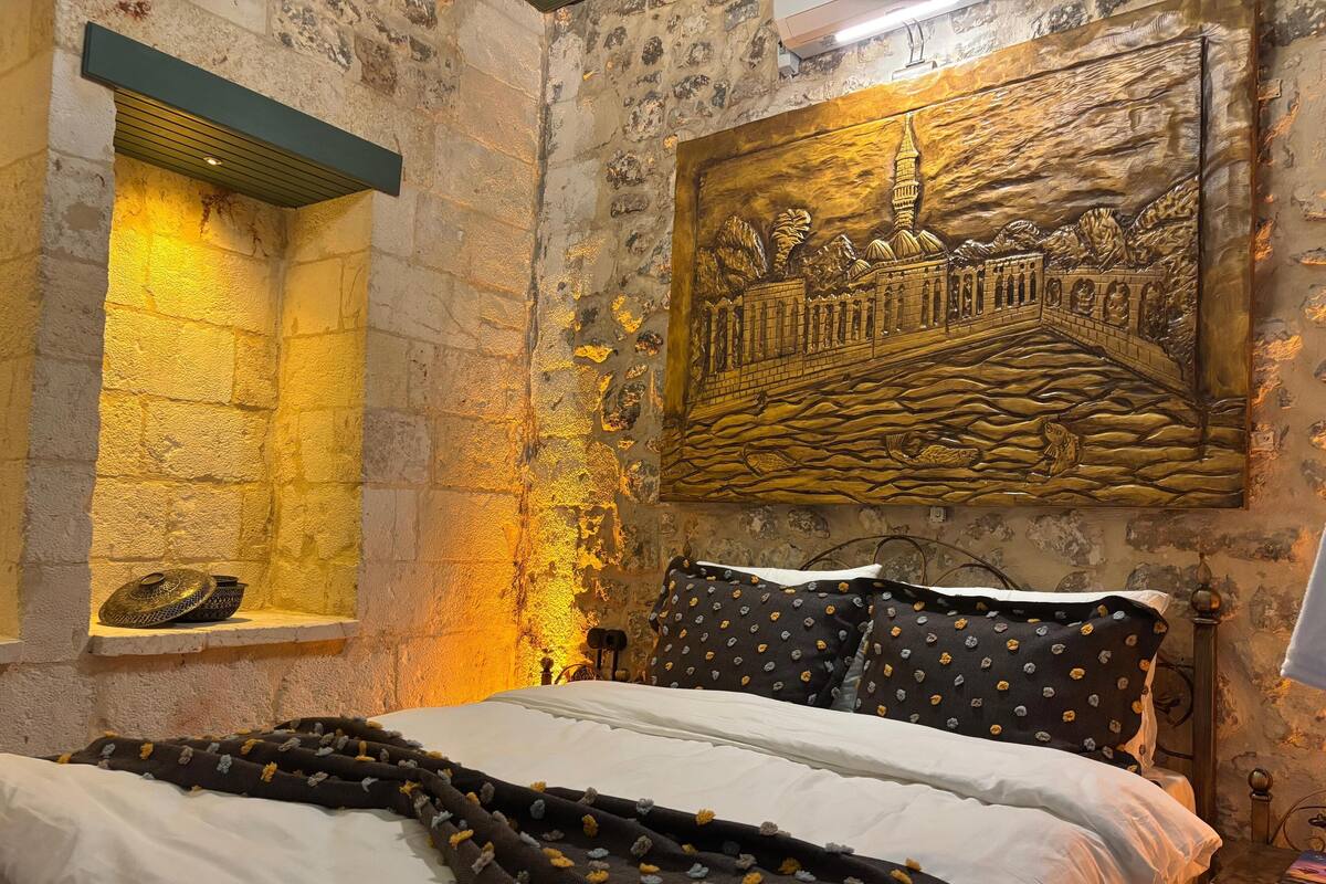 Une chambre avec un mur en pierre, un lit avec une literie blanche et à motifs, une étagère avec des objets décoratifs et un tableau accroché au mur.