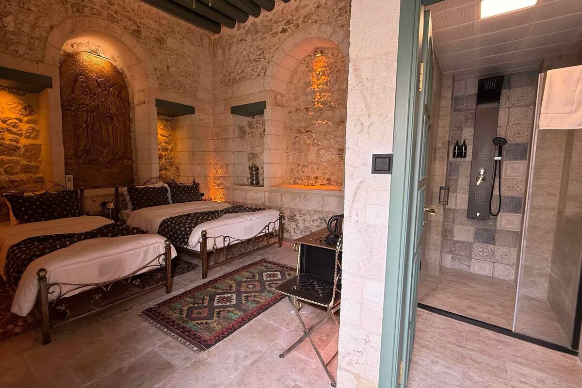 Une pièce aux murs de pierre, avec deux lits dotés de draps sombres, un tapis à motifs et une douche moderne.
