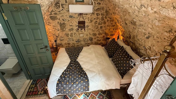 Una stanza con parete in pietra, un letto con copriletto decorato, una porta verde e un piccolo ripiano bianco.