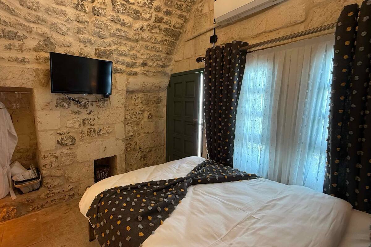 Une chambre avec un mur en pierre, un téléviseur à écran plat fixé au-dessus d’une cheminée, un lit recouvert d’une couverture blanche à motifs et une fenêtre avec des rideaux foncés.