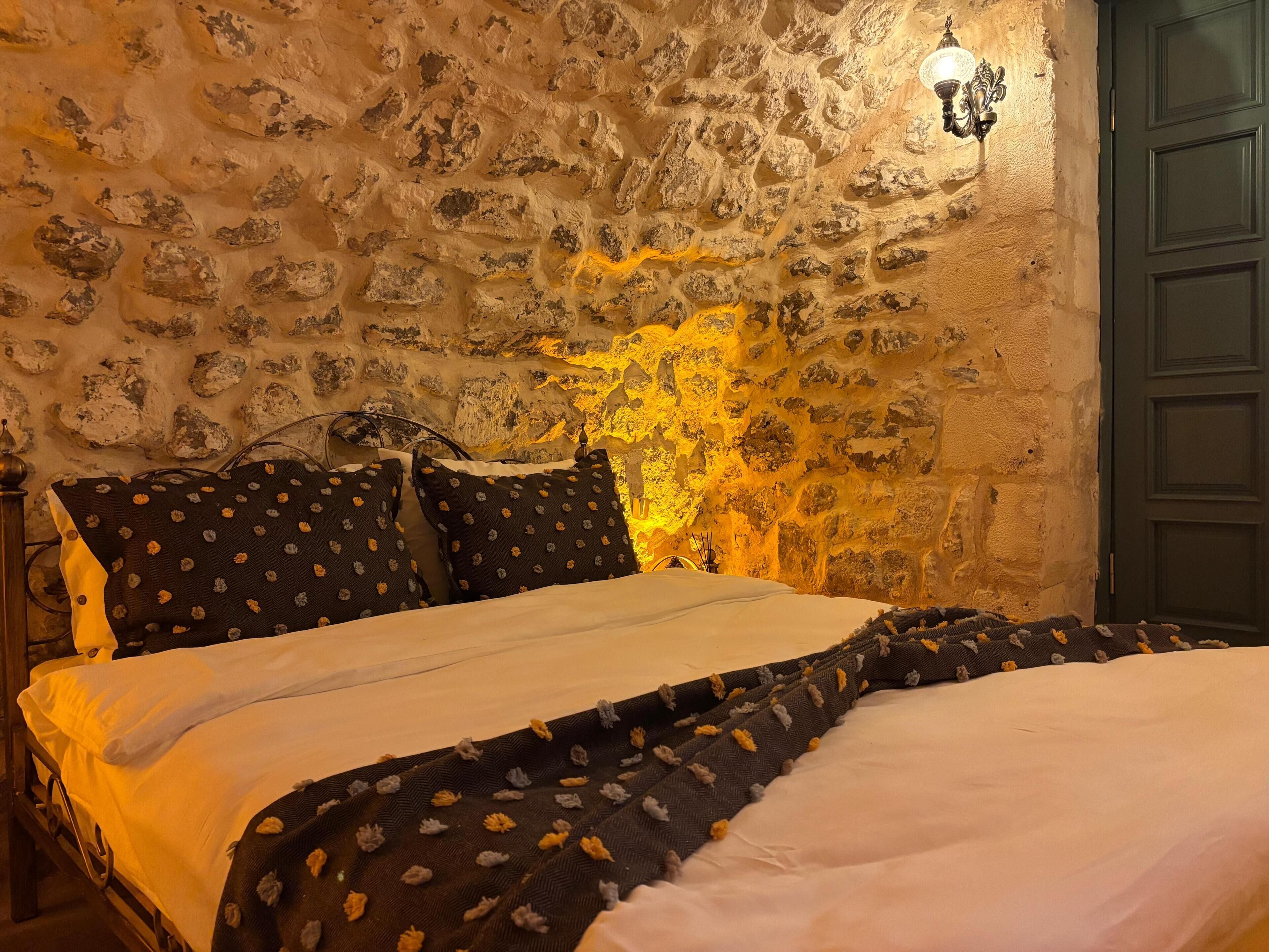 Quarto com parede de pedra, cama com colcha estampada escura e porta verde.