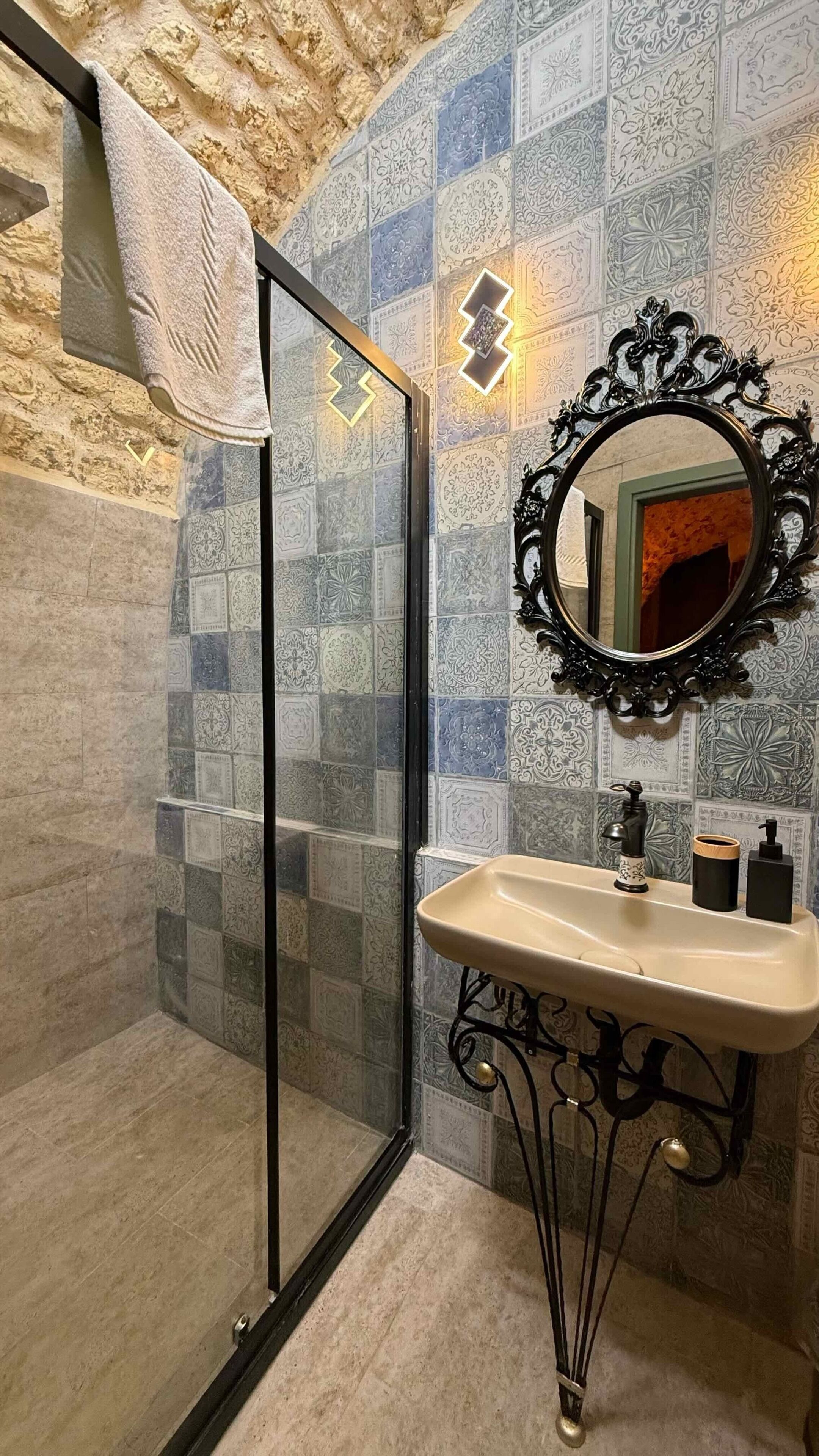 Un baño con mampara de vidrio, un lavamanos con un espejo decorativo y azulejos estampados en las paredes.