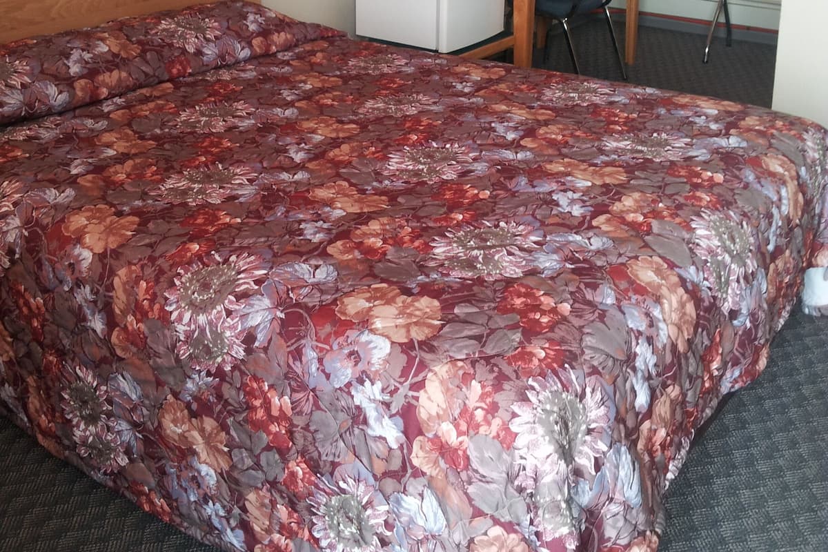 A hotel room with a large floral-patterned bed, a desk with a chair, a small refrigerator, and a mirror on the wall.