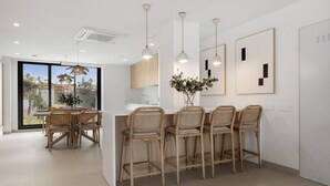 A modern kitchen with a central island, wicker chairs, pendant lights, and a view through large windows. - Venían Views 2BR Selwo Estepona (Málaga)