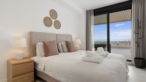A modern bedroom with a large bed, two bedside tables, and a view of the sea through a large window. - Venían Views 2BR Selwo Estepona (Málaga)