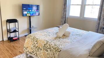 Ein Schlafzimmer mit einem Bett, einem an der Wand montierten Fernseher, einer kleinen Ablage und einem Fenster mit Vorhängen.