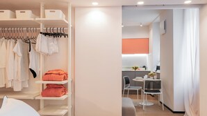 Une chambre d’hôtel avec un lit, une armoire où sont suspendus des vêtements, un coin salon avec une chaise et une table, et une petite cuisine en arrière-plan.