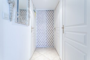 A hallway with white walls, a mirrored wall, a door with a white panel, and a tiled floor.