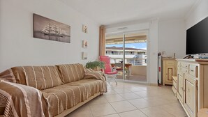 A living room with a sofa, a TV, a balcony with a view, and a dining area. - Apartment 'Petit Paradis' with Shared Pool, Wi-Fi, and Air Conditioning (Cavalaire-sur-Mer)