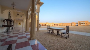 Une cour avec un sol à carreaux, une table entourée de chaises et, au fond, un bâtiment orné d’arches.