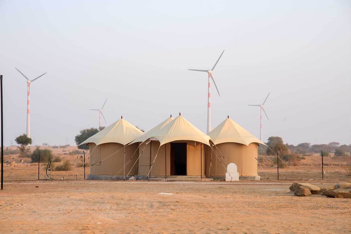 A desert landscape with three tents and wind turbines in the background.