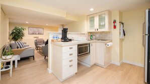 A modern kitchen with white cabinetry, a built-in oven, and a coffee machine. There is a dining area with a gray sofa and a small table. - 2BR Trafalgar Retreat near UBC & Pacific Spirit (Vancouver)