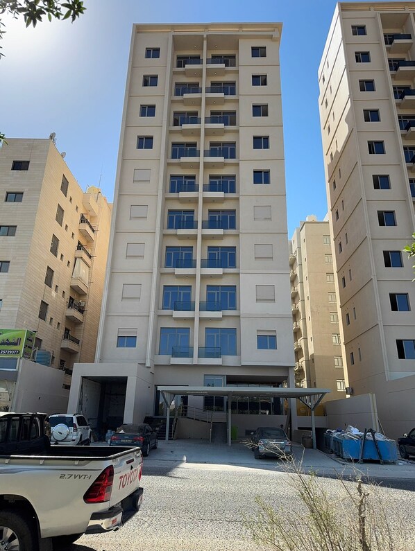 A multi-story residential building with balconies, a covered entrance, and a parking area with vehicles.
