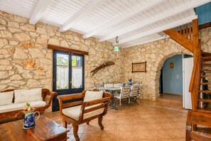 Una habitación con paredes de piedra, un banco de madera, una mesa con sillas, una lámpara colgante y una escalera.