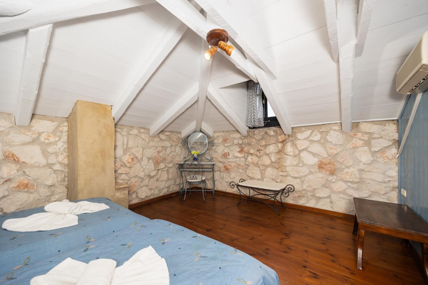 A bedroom with a sloped ceiling, stone walls, a bed with a blue bedspread, a bedside table, a mirror, and a hanging light fixture.