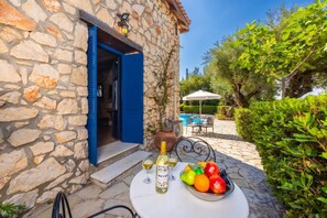 Un bâtiment en pierre avec une porte bleue, une table avec une bouteille de vin et un saladier de fruits, et une piscine en arrière-plan.