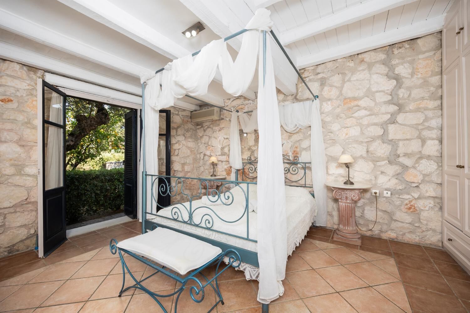 A four-poster canopy bed with a white canopy, a small table with a lamp, and a stone wall.