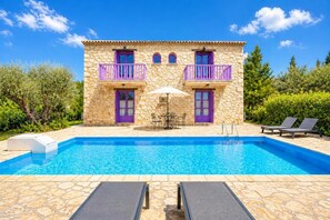 A stone building with a swimming pool, two balconies with purple railings, and outdoor seating.
