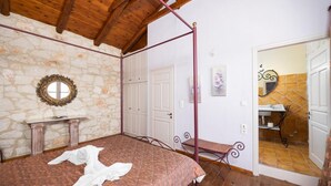 A bedroom with a stone wall, a bed, a wooden ceiling, a mirror, and a bathroom visible through an open door. - Villa Carolina - Two Bedroom Villa, Sleeps 5 (Agios Dimitrios)