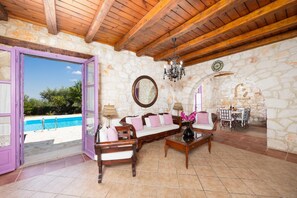 Habitación con pared de piedra, vigas de madera en el techo, una piscina visible a través de una puerta, un sofá, una mesa con sillas y una araña.