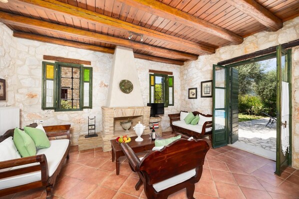 A rustic interior with stone walls, wooden furniture, a fireplace, and a view of greenery outside.