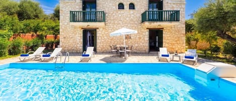 Un edificio de piedra con piscina, zona de estar al aire libre y cielo azul despejado.